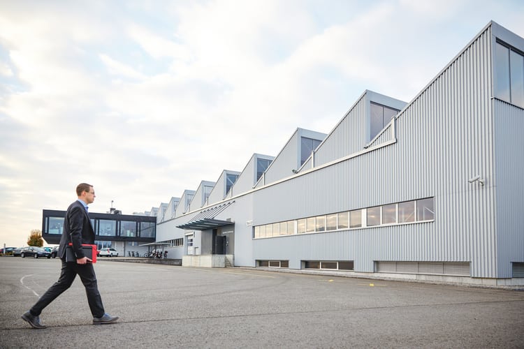Ein Mann läuft auf ein Industriegebäude zu. Er hält eine rote Mappe unter dem Arm. 