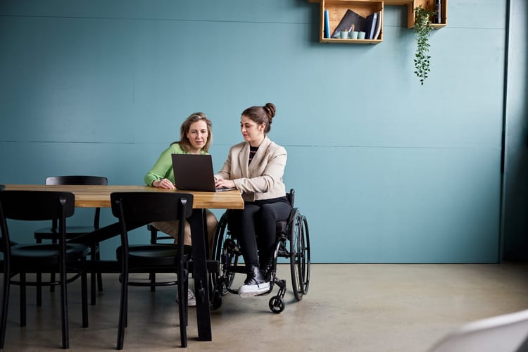 Zwei Frauen sitzen an einem Holztisch und schauen sich etwas auf einem Laptop an. Die Frau rechts sitzt im Rollstuhl. 