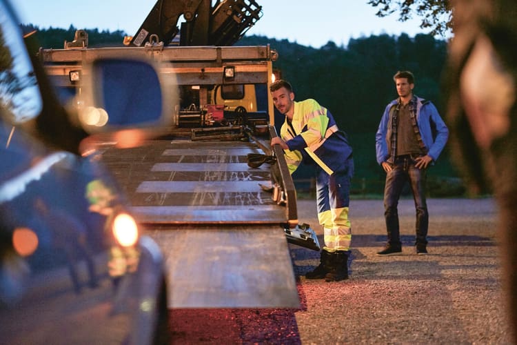 Ein Pannenhelfer lädt mit der Seilwinde ein offensichtlich beschädigtes Auto auf die Ladebrücke des Pannenfahrzeugs. Der Besitzer des Autos steht neben ihm und schaut zu. 