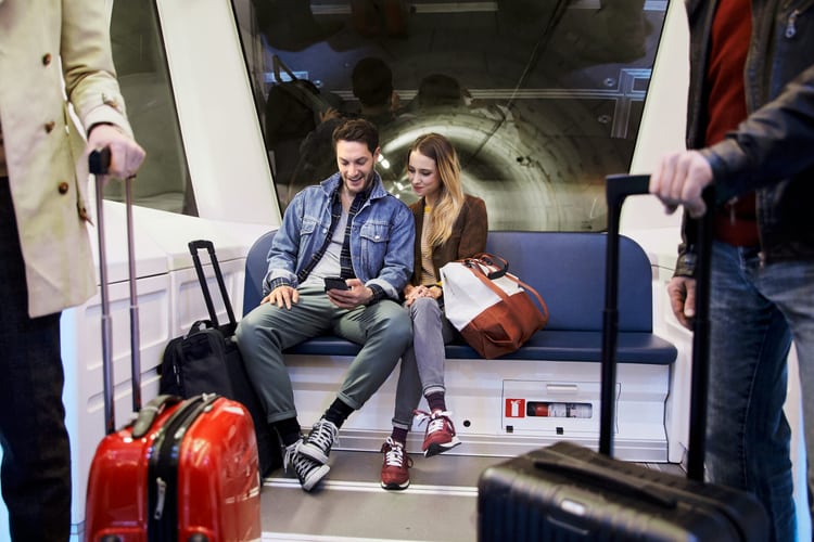 Ein Mann und eine Frau sitzen im hintersten Wagen einer U-Bahn, sie schauen auf das Handy, dass der Mann in der Hand hält. Vor ihnen stehen Personen mit Reisegepäck. Symbolbild für das Thema Datenschutz für kleine Unternehmen.