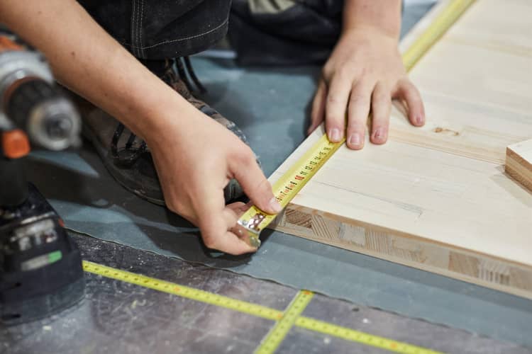 Eine Person vermisst ein Stück Holz, das am Boden liegt. Sichtbar sind nur die Hände der Person. Unscharf – etwas im Vordergrund – sieht man eine Bohrmaschine. 