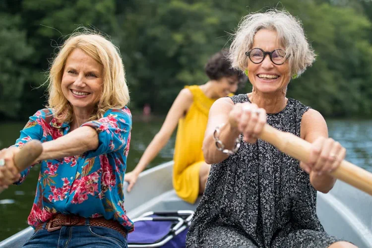 Zwei rund 50-jährige Frauen rudern ein Boot. Eine dritte Frau in einem gelben Kleid sitzt als Passagierin an der Spitze des Boots. 