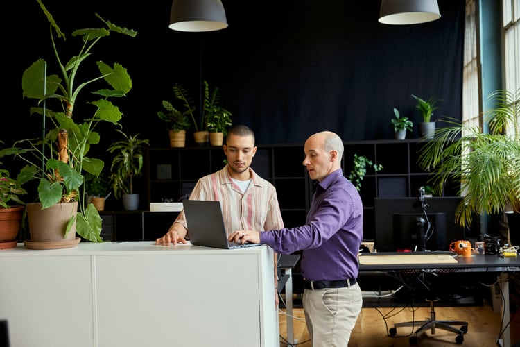 Deux collègues de travail discutent debout et regardent le laptop placé devant eux sur un séparateur de pièce. 