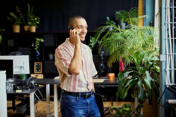 Un uomo sta telefonando in piedi in un ufficio, in cui ci sono tante piante. Sullo sfondo si vede una scrivania. 