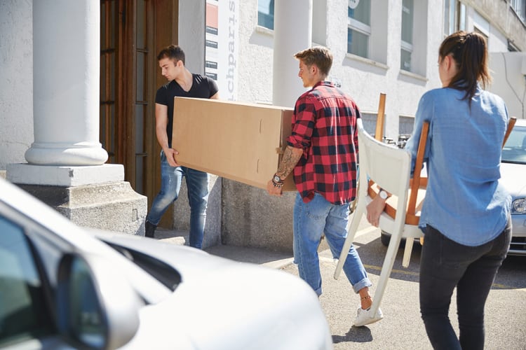 Zwei Männer und eine Frau tragen einen Schrank und ein paar Stühle zusammen in das neue Wohnhaus. 
