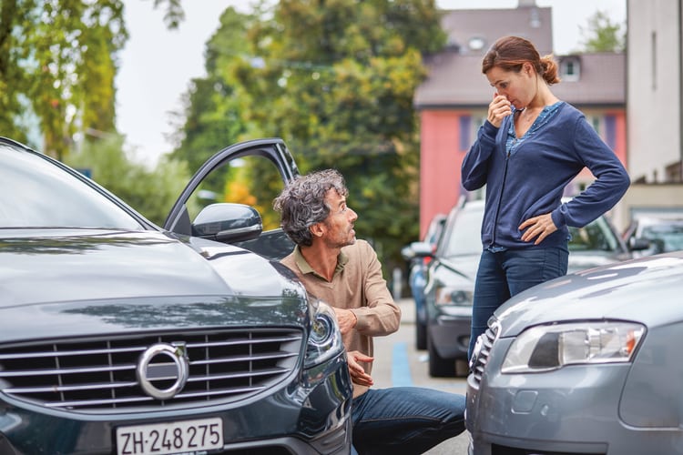Un homme est agenouillé à côté de sa voiture et parle à une femme qui se tient à côté de sa propre voiture et l’écoute. Il semble évaluer un dommage dû à une collision entre les deux véhicules. 
