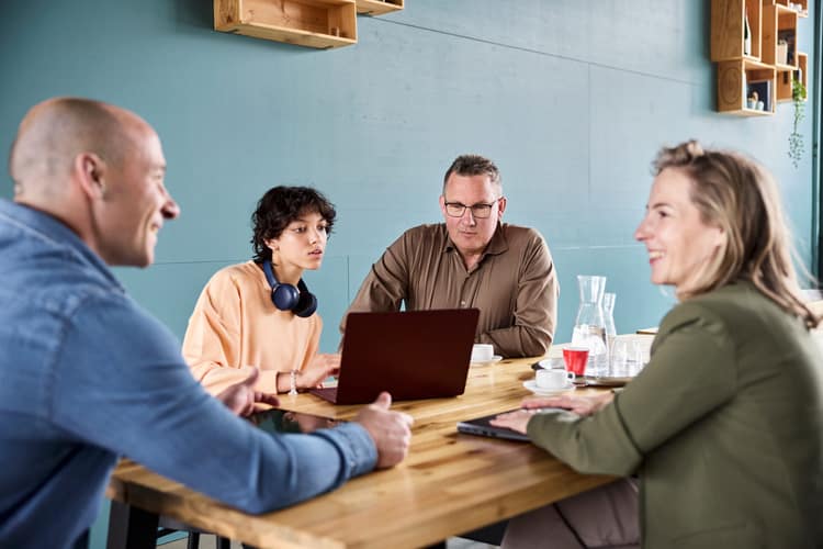 Due uomini e due donne siedono a un tavolo. Mentre due di loro parlano, gli altri due guardano lo schermo di un computer portatile. 
