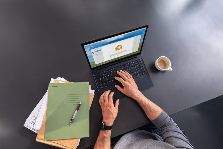 Person bedient einen Laptop auf dem Tisch. Daneben liegen ein Stapel Papier und eine Tasse Kaffee. 