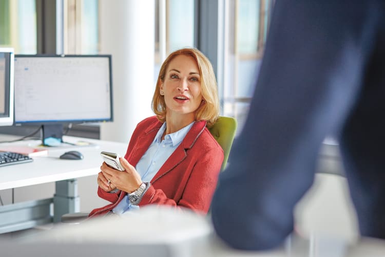 Frau sitzt am Arbeitsplatz mit dem Handy in der Hand und blickt zum Kollegen