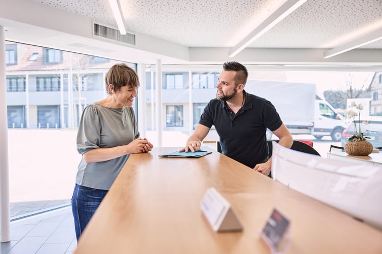 Ein Mann hinter einem Kundentresen gibt einer Frau eine blaue Arbeitsmappe. Beide lachen. 
