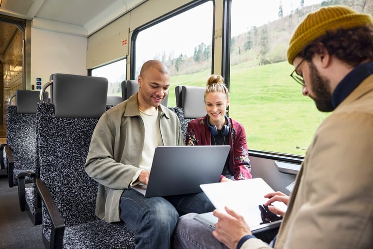 Eine Gruppe junger Menschen sitzt im Zug, alle arbeiten an einem Laptop oder Tablet. 