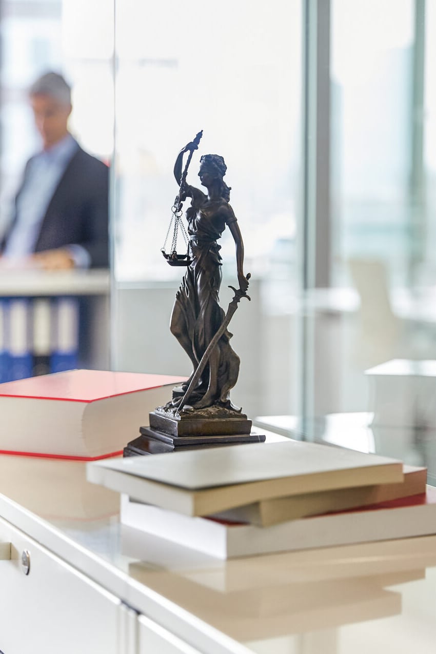 In einem Grossraumbüro steht im scharfen Vordergrund auf einem Korpus eine kleine Justitia-Statue mit Waage und Schwert. Im unscharfen Hintergrund diskutieren zwei Männer miteinander. 