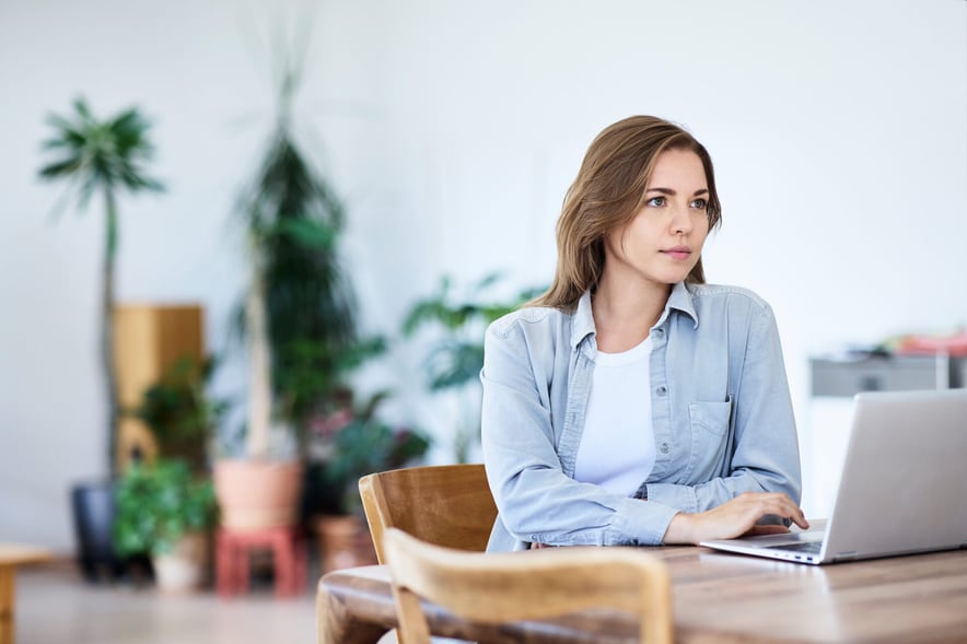 Une jeune femme est assise à une table de salon devant son ordinateur portable, le regard plongé dans ses pensées.