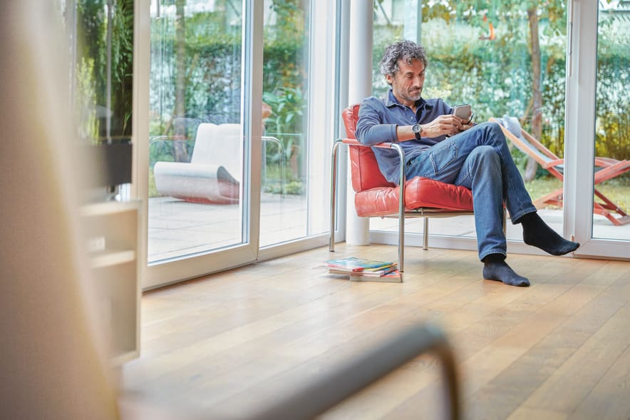 Un uomo è seduto rilassato su una sedia con cuscini rossi in salotto e legge sul suo cellulare.