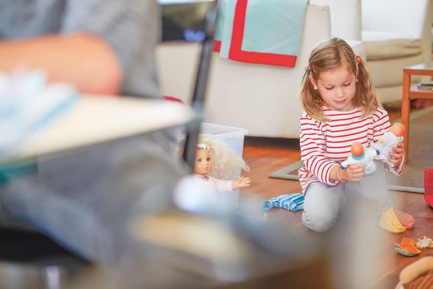 Ein Kind spielt im Hintergrund am Boden. Im Vordergrund ist ein Ellbogen einer Frau zu sehen, die an einem Tisch am Laptop sitzt vor einem offenen Ordner.