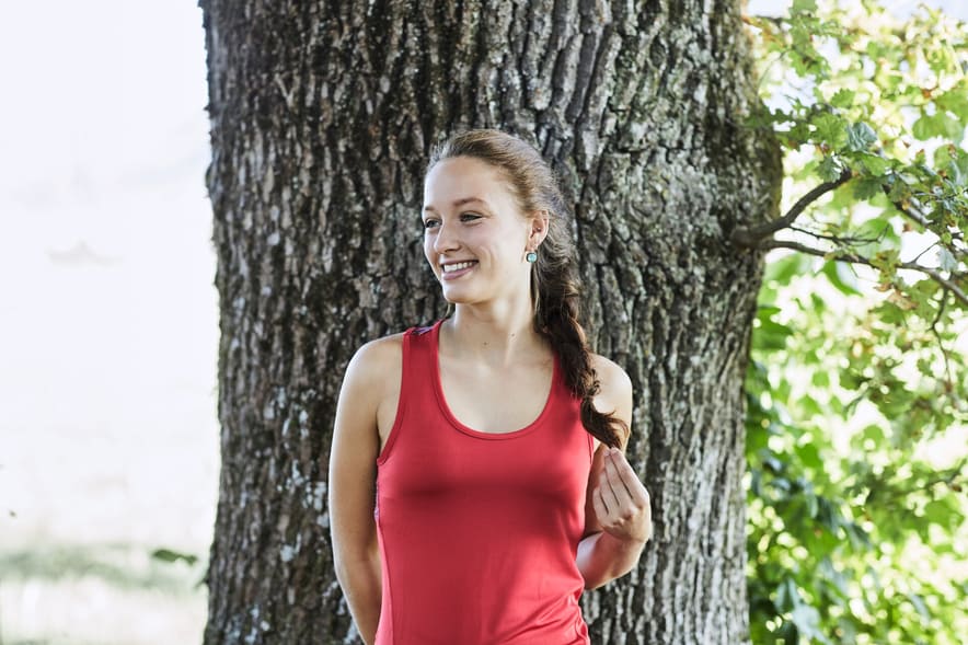 Une jeune femme avec un débardeur rouge et des cheveux bruns tressés se tient devant un tronc d’arbre et regarde sur le côté en souriant.