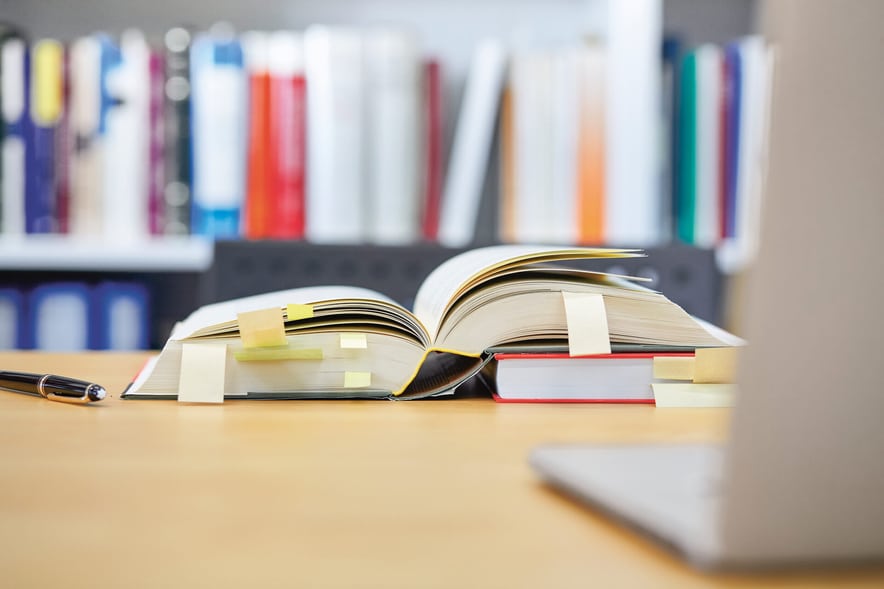 Un libro aperto su un tavolo, molte pagine sono segnate, sullo sfondo c’è una libreria piena.