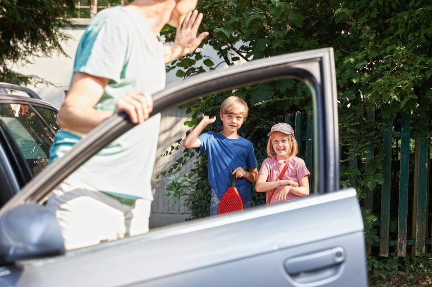 Un père se tient dans la portière ouverte de sa voiture argentée et fait signe à ses deux enfants qui se tiennent devant la maison avec des sacs.