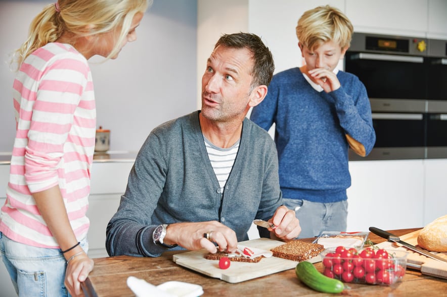 Un homme prépare des petits pains à la table de la cuisine, tandis que sa fille et son fils en âge d’aller à l’école primaire le regardent. 