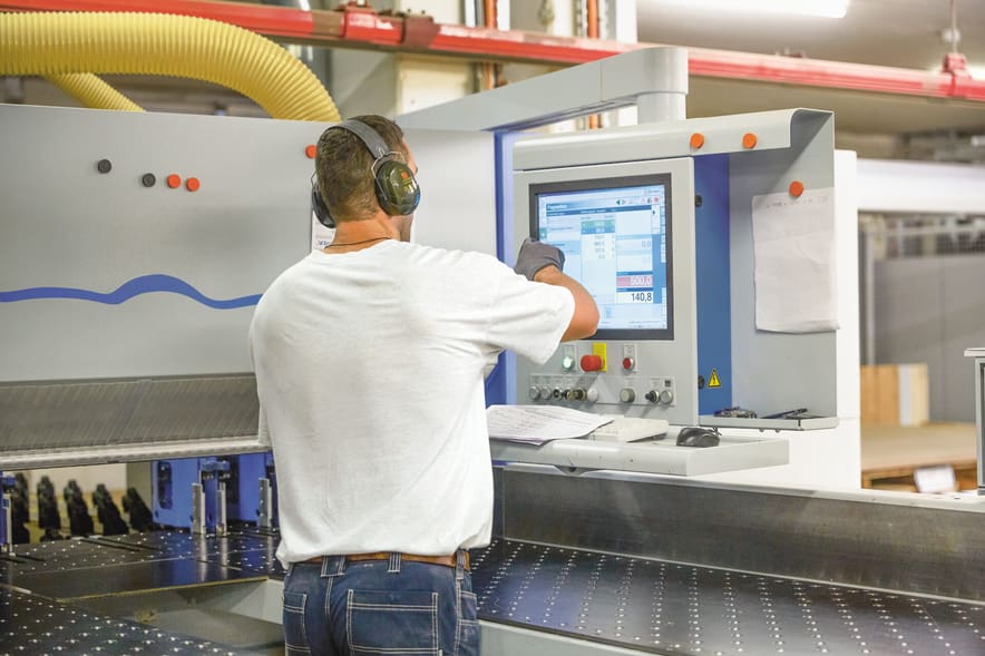 Un operaio indossa una protezione acustica e si trova di fronte a una macchina di produzione e comanda il pannello di controllo.