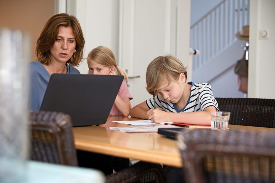 Une mère est assise à table avec ses enfants. La mère est sur l’ordinateur portable pendant que les enfants dessinent quelque chose.