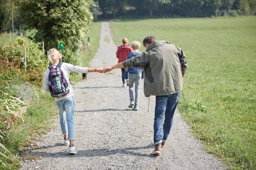 Eine Familie wandert auf einem Feldweg einem Waldrand entlang. Der Vater und ein Kind halten sich an einer Hand.