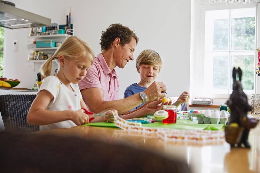 Un padre è seduto al tavolo della cucina con la figlia e il figlio, e sta tingendo le uova con loro.