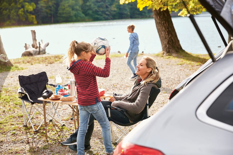 Una donna è seduta su una sedia da campeggio, su un tavolo da campeggio pieno di Piquenique, davanti al portellone aperto del bagagliaio di un’automobile. Sta parlando con la figlia, che tiene in mano un pallone da calcio, e suo figlio gioca sullo sfondo.