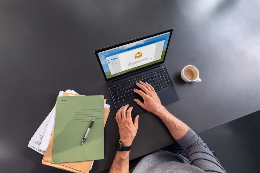 Person bedient einen Laptop auf dem Tisch. Daneben liegen ein Stapel Papier und eine Tasse Kaffee. 