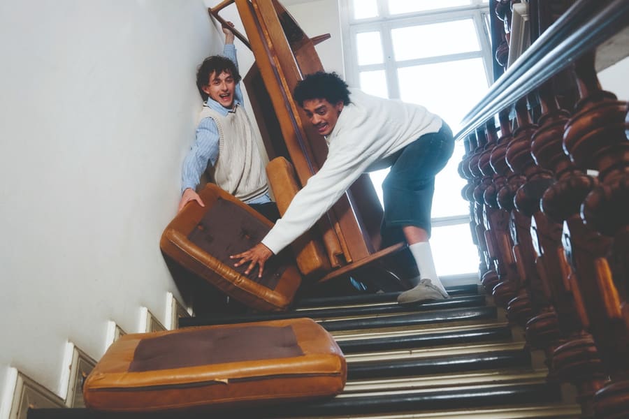 Zwei junge Männer manövrieren mit sichtlicher Mühe ein Sofa eine enge Treppe hinunter.