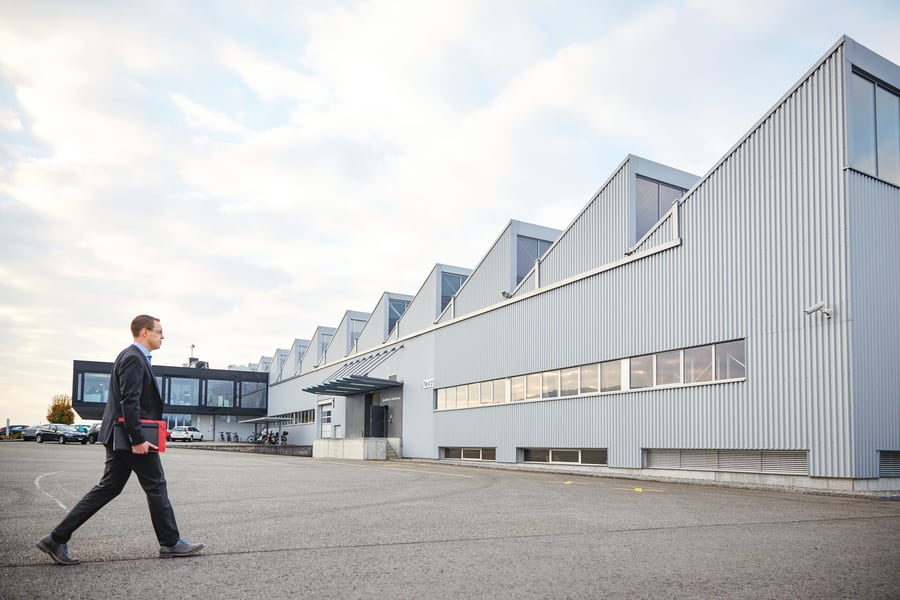 Ein Mann läuft auf ein Industriegebäude zu. Er hält eine rote Mappe unter dem Arm. 