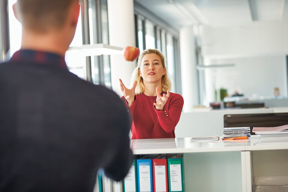 In un ufficio ci sono una donna e un uomo l’uno di fronte all’altro e l’uomo lancia una mela alla donna.