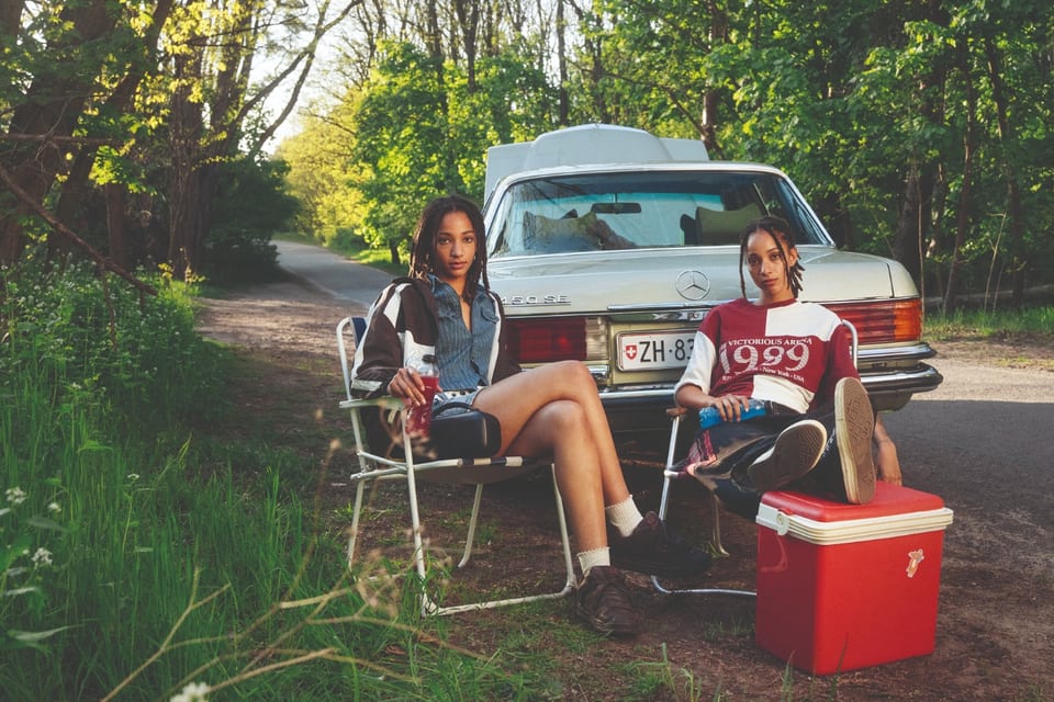  Deux jeunes femmes sont assises détendues devant une voiture de collection au capot ouvert abandonnée à l’orée d’une forêt. Elles tombent en panne sur une route de campagne.