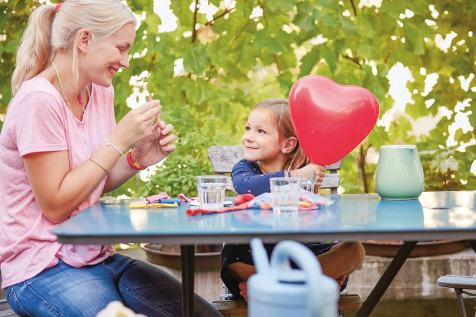 Invalidenrente für Kinder: Eine junge Frau und ein Mädchen sitzen an einem Gartentisch vor einem Baum und strahlen einander an. Das Mädchen hält einen roten Herzballon in der Hand. Auf dem Tisch hat es Trinkgläser, einen Wasserkrug und nicht aufgeblasene Ballone. 