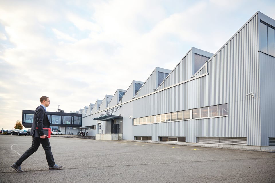 Ein Mann läuft auf ein Industriegebäude zu. Er hält eine rote Mappe unter dem Arm.