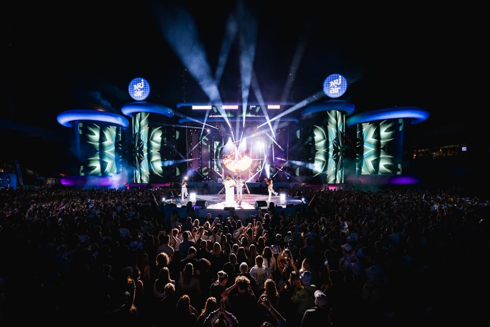 NRJ Air Bühne im Dunkeln mit Lichtshow und Sänger. Davor das Publikum von hinten.