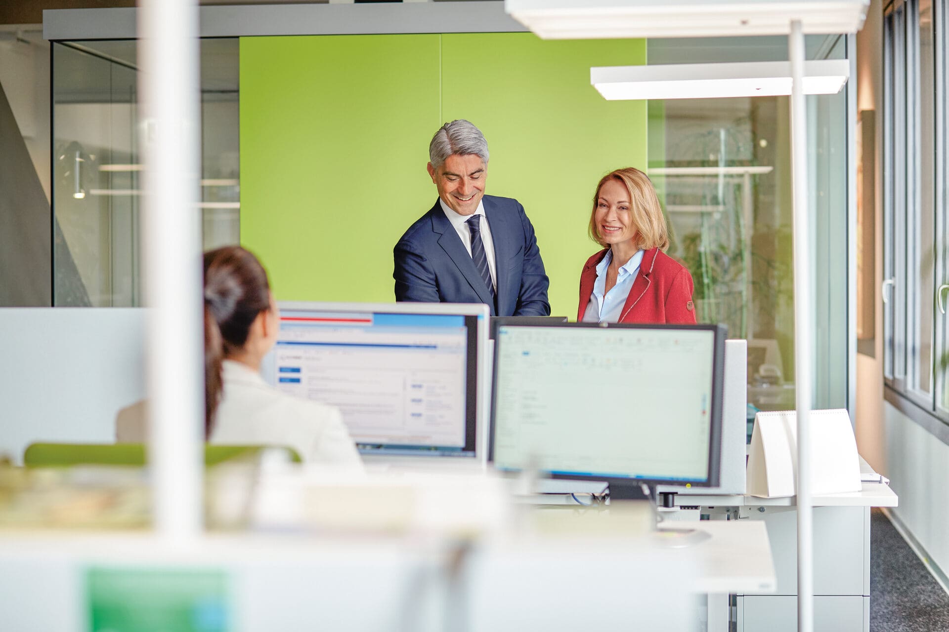 Eine Frau sitzt in einem Grossraumbüro und arbeitet am Computer. Gegenüber von ihr stehen eine Frau und ein Mann, die mit ihr sprechen. 