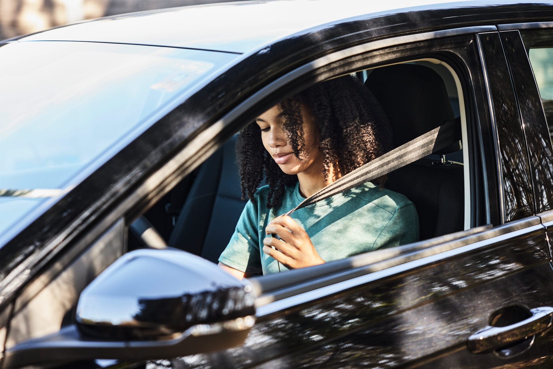 Una giovane donna siede al volante di un’auto nera e si allaccia le cinture di sicurezza. Il sole e un albero si riflettono nell’auto. 