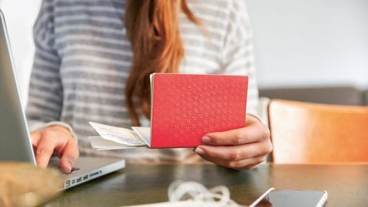 Una donna tiene il passaporto svizzero con la sinistra e con la destra tocca sulla tastiera del laptop.
