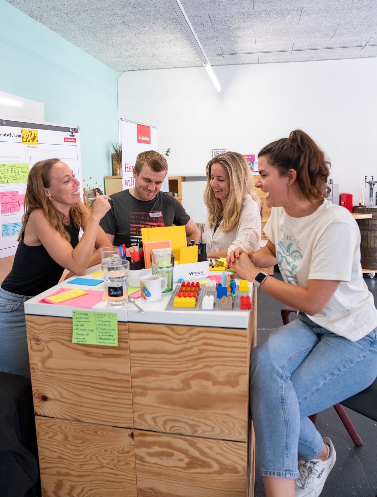 Vier junge Erwachsene sitzen an einem hohen Holztisch, lachen und arbeiten kreativ mit bunten Karten, Stiften und LEGO-Steinen – eine Szene aus einem partizipativen Workshop oder Ideensprint.
