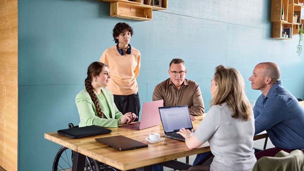 Eine Gruppe von 5 Personen bespricht etwas am Tisch mithilfe von Laptops.