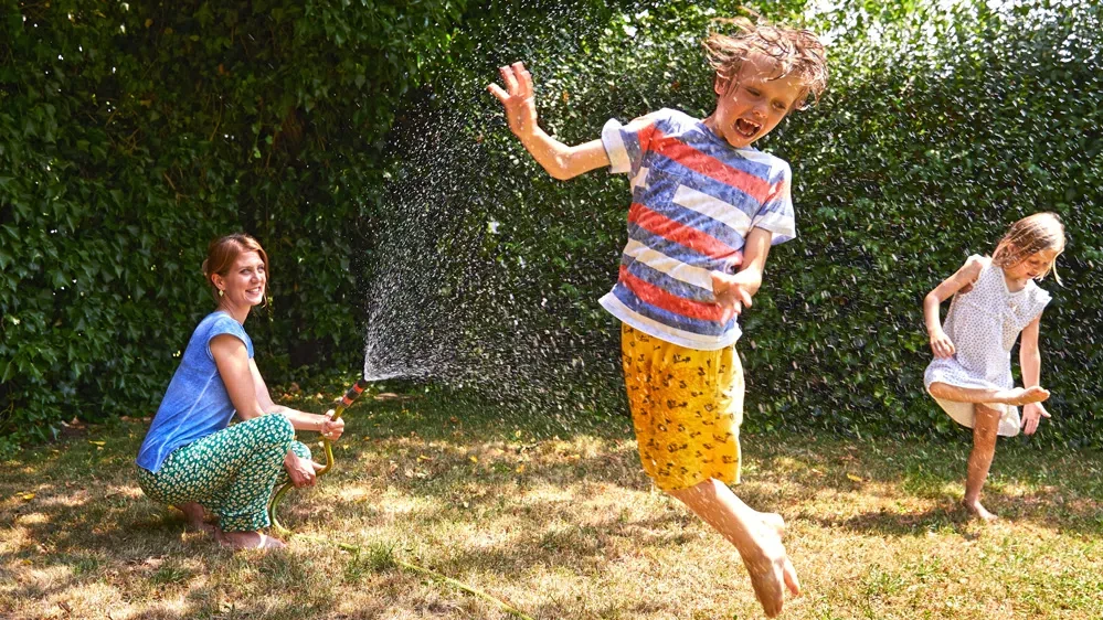 Eine Mutter kauert im Garten und bespritzt Ihre zwei Kinder mit einem Wasserschlauch. 