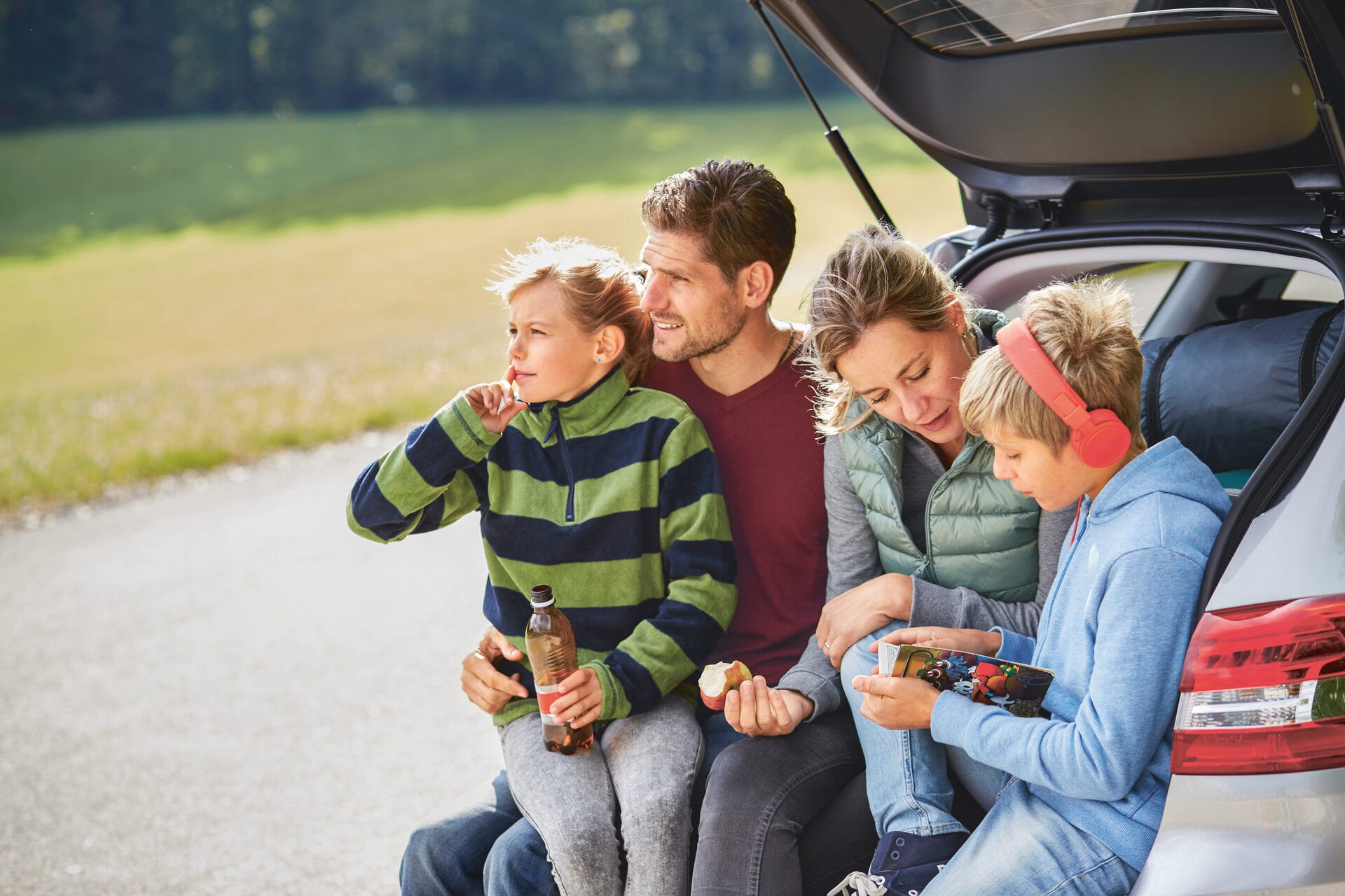 Familie mit Mutter, Vater, Tochter und Sohn sitzt bei offenen Kofferraumdeckel im Heck ihres Fahrzeugs.