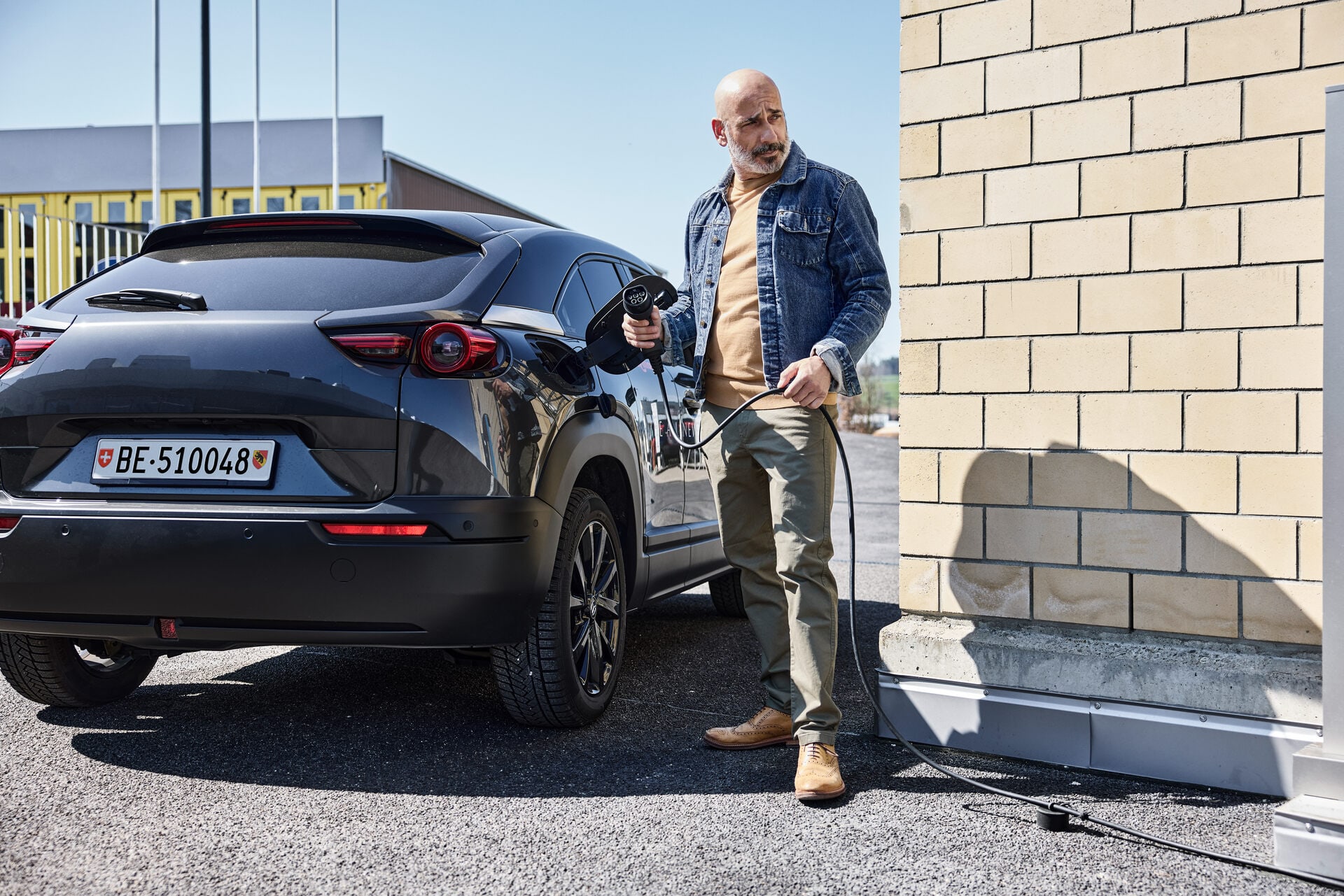 Un homme d’âge moyen, chauve et vêtu d’une veste en jean, se tient prêt à faire le plein avec une station de recharge à côté d’une voiture électrique noire.