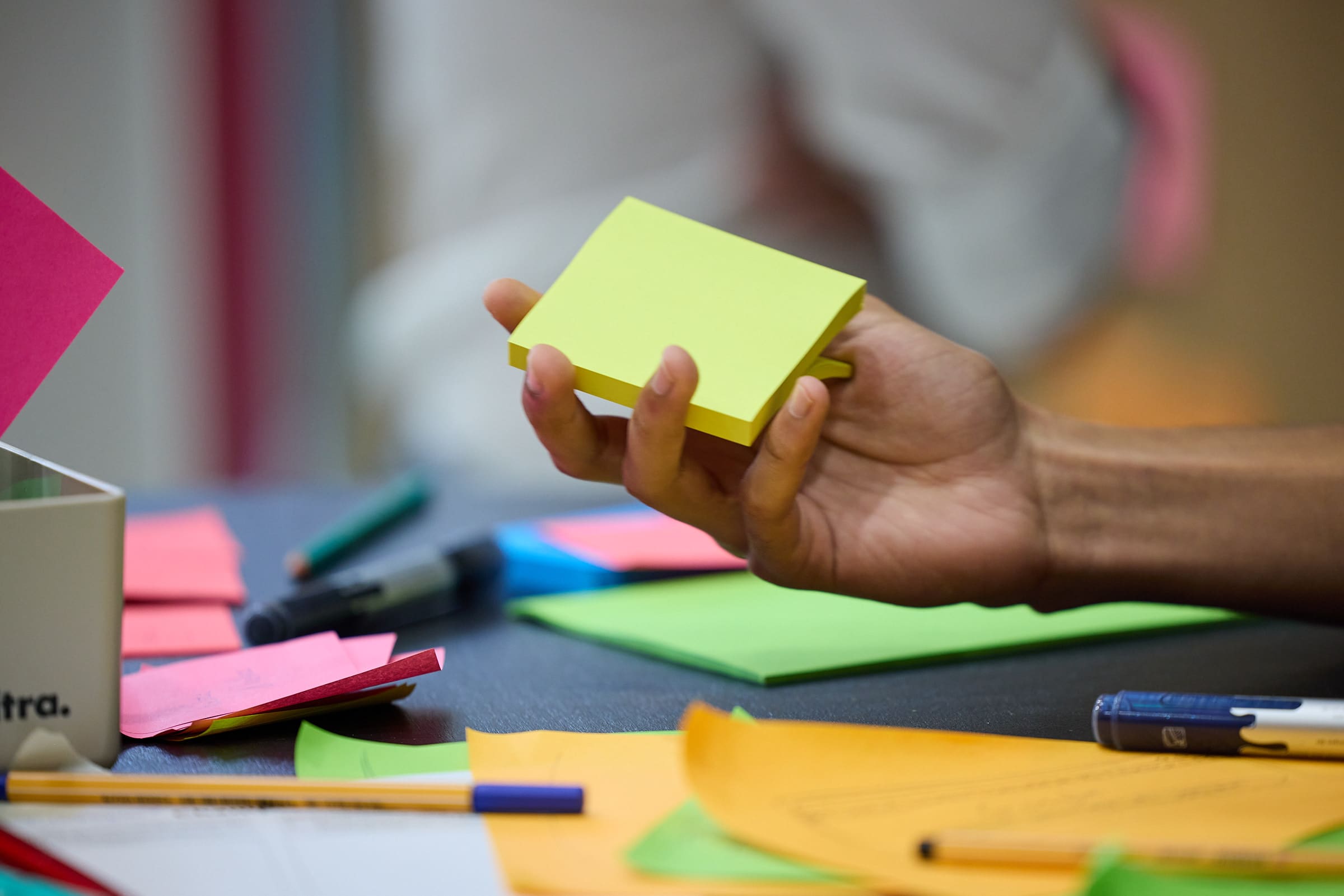 Auf einem Tisch liegen viele Post-its. Jemand hält einen hellgrünen Post-it-Block in der Hand. Sichtbar ist nur die Hand der Person.