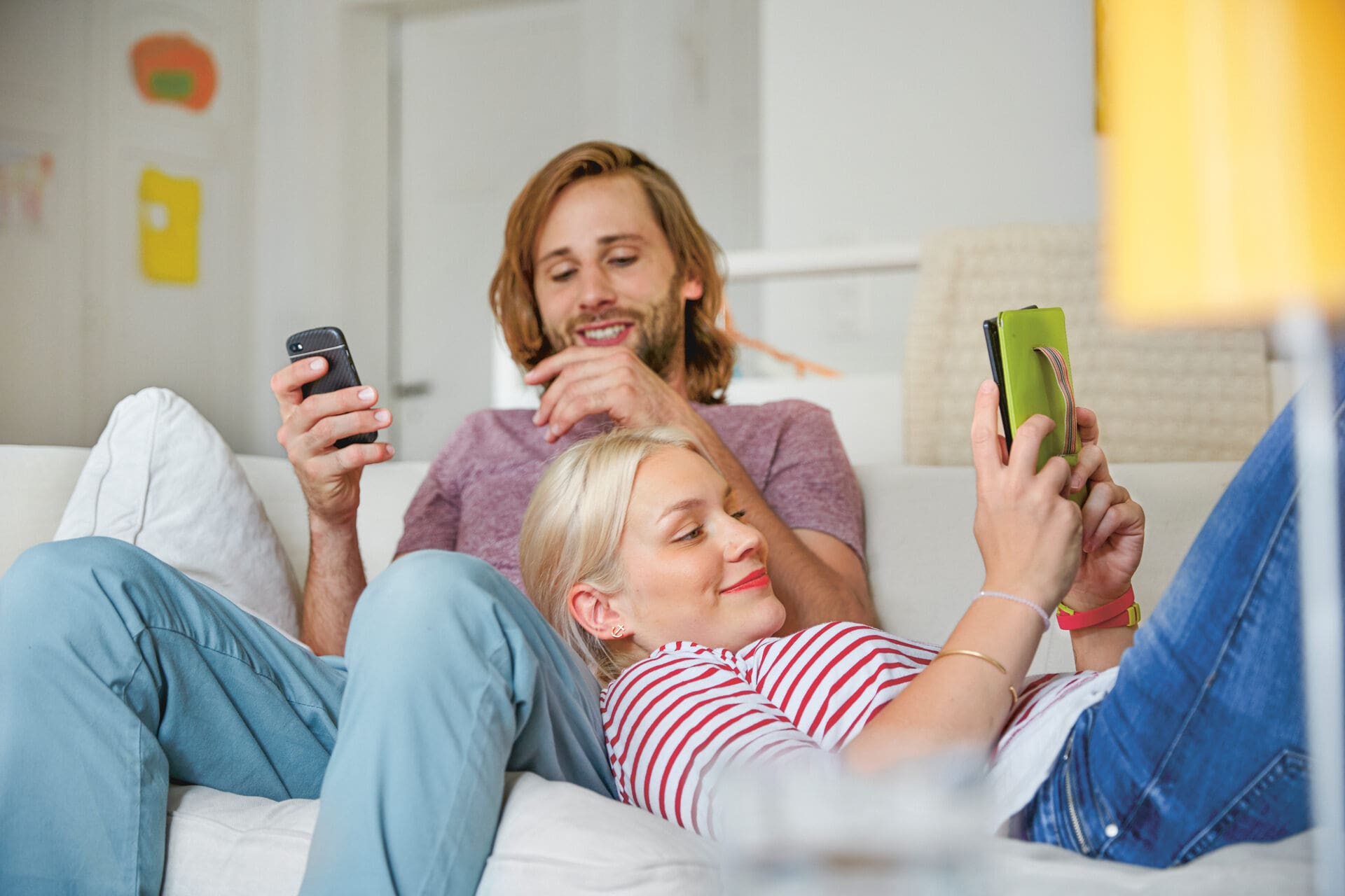 Ein Paar liegt auf dem Sofa in einem Wohnzimmer. Der Mann sitzt und die Frau liegt, ihren Kopf auf das Bein ihres Partners gelegt. Beide haben ein Handy in der Hand.