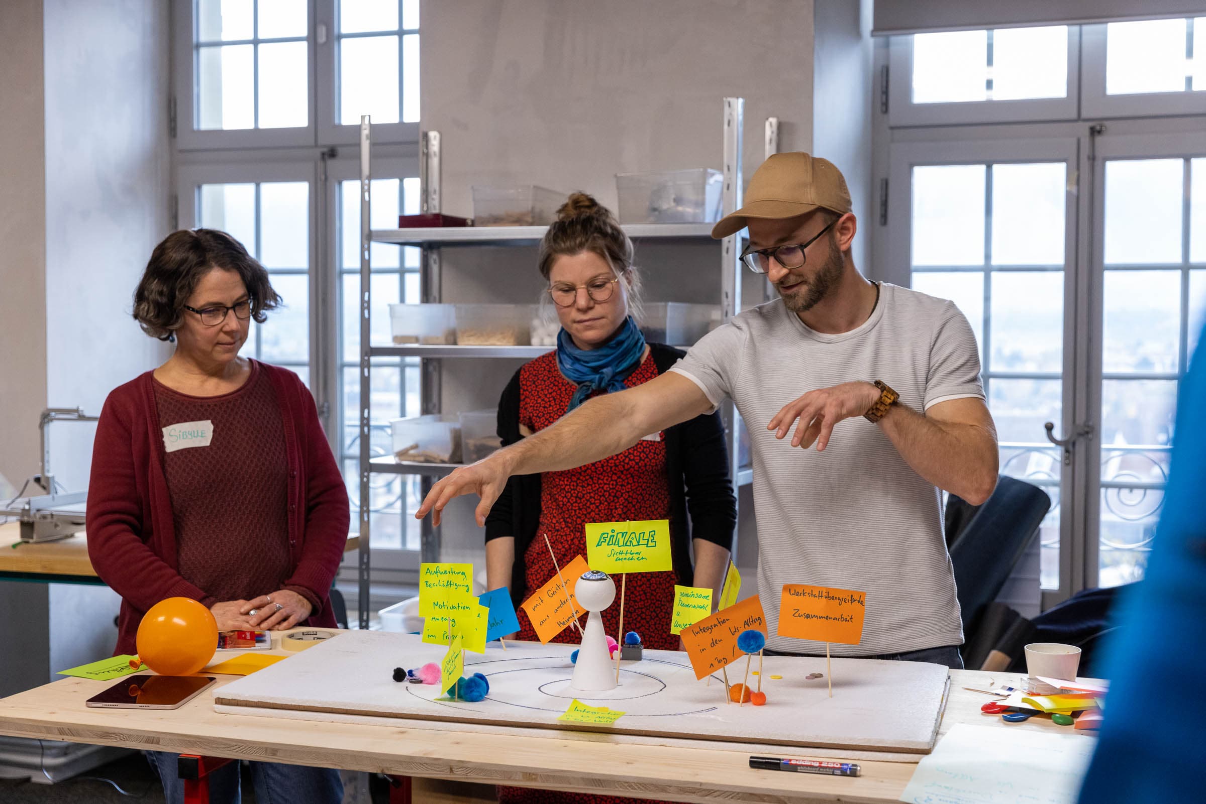 Eine Gruppe von Teilnehmer:innen bespricht die erarbeiteten Resultate. Zwei Frauen und ein Mann stehen rund um einen Tisch, auf welchem sie das erarbeitete Modell präsentieren.