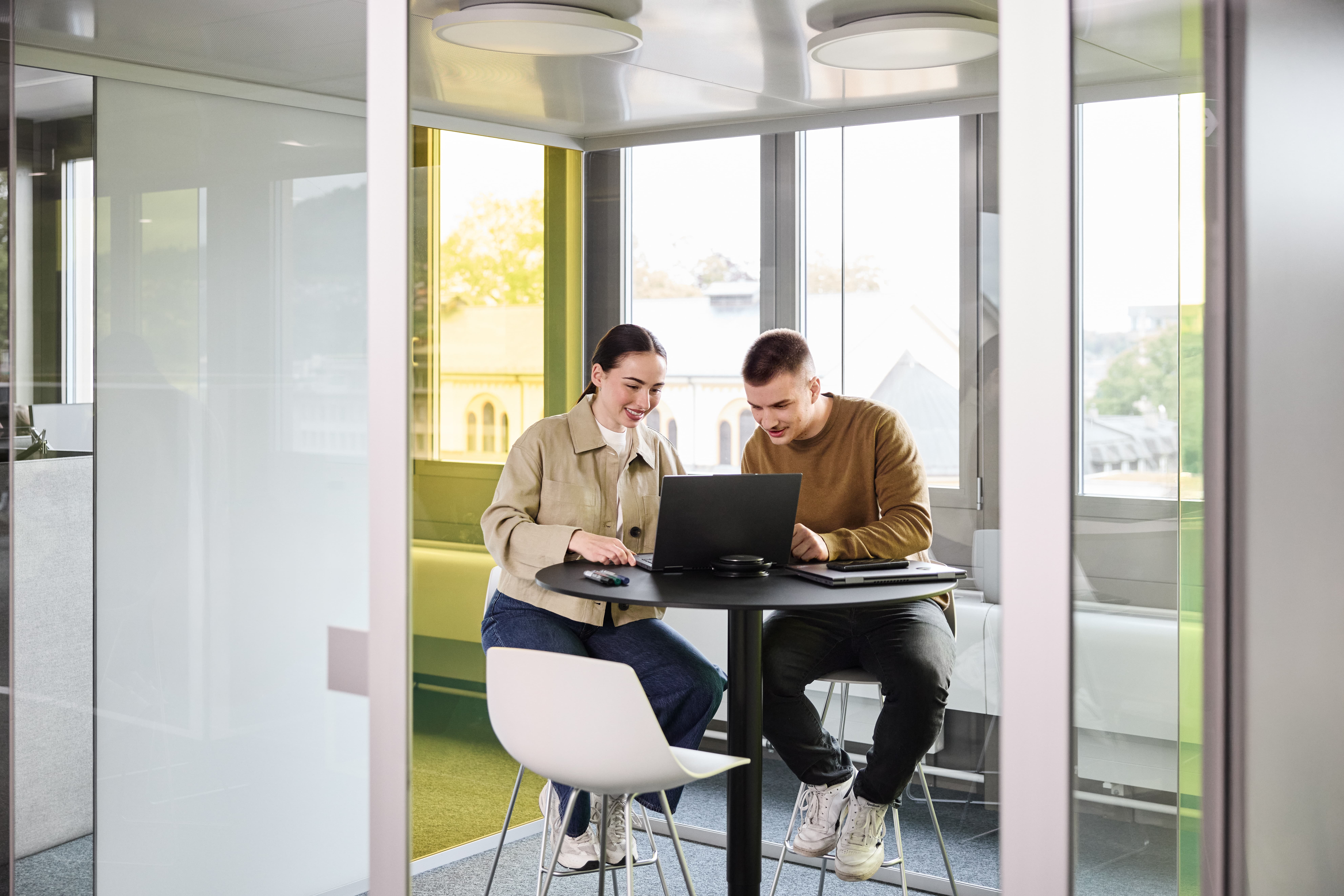 Ein Mann und eine Frau unterhalten sich im Stehen. Eine weitere Frau steht daneben und arbeitet auf ihrem Laptop, der auf einem Stehtisch platziert ist.