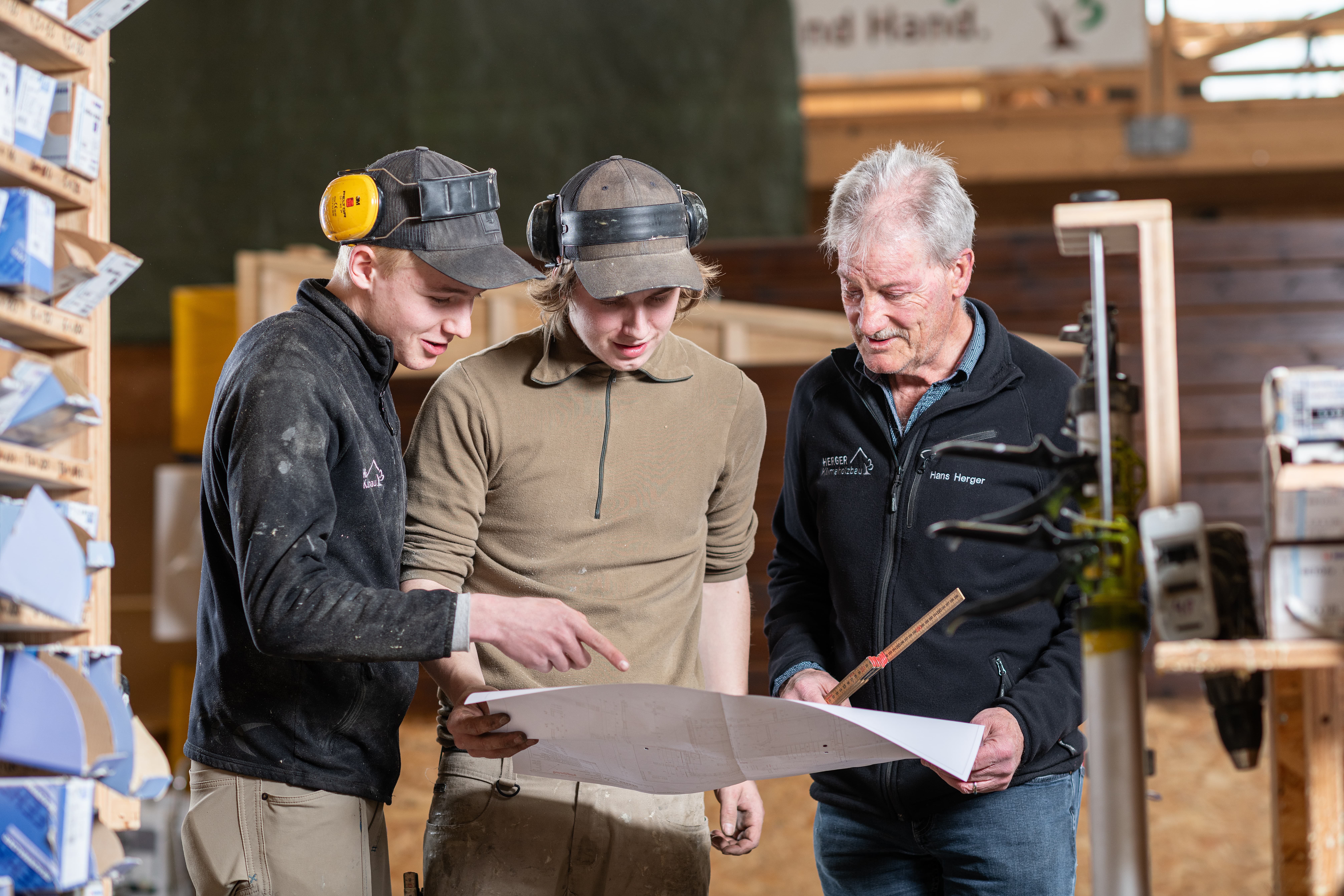 Drei Männer stehen in einer Holzwerkstatt und schauen sich gemeinsam einen Plan an.