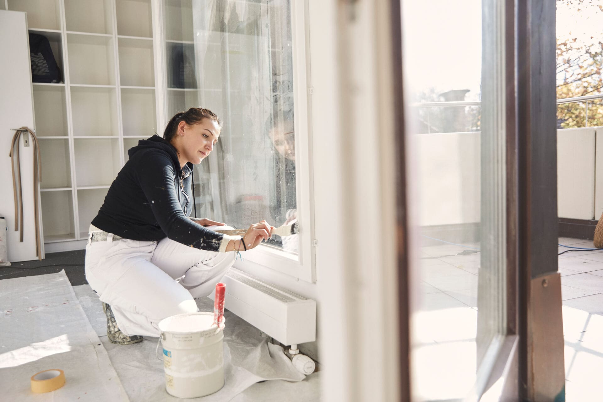 Eine Malerin streicht einen Fensterrahmen in einem Neubau neben einer offenen Balkontür.